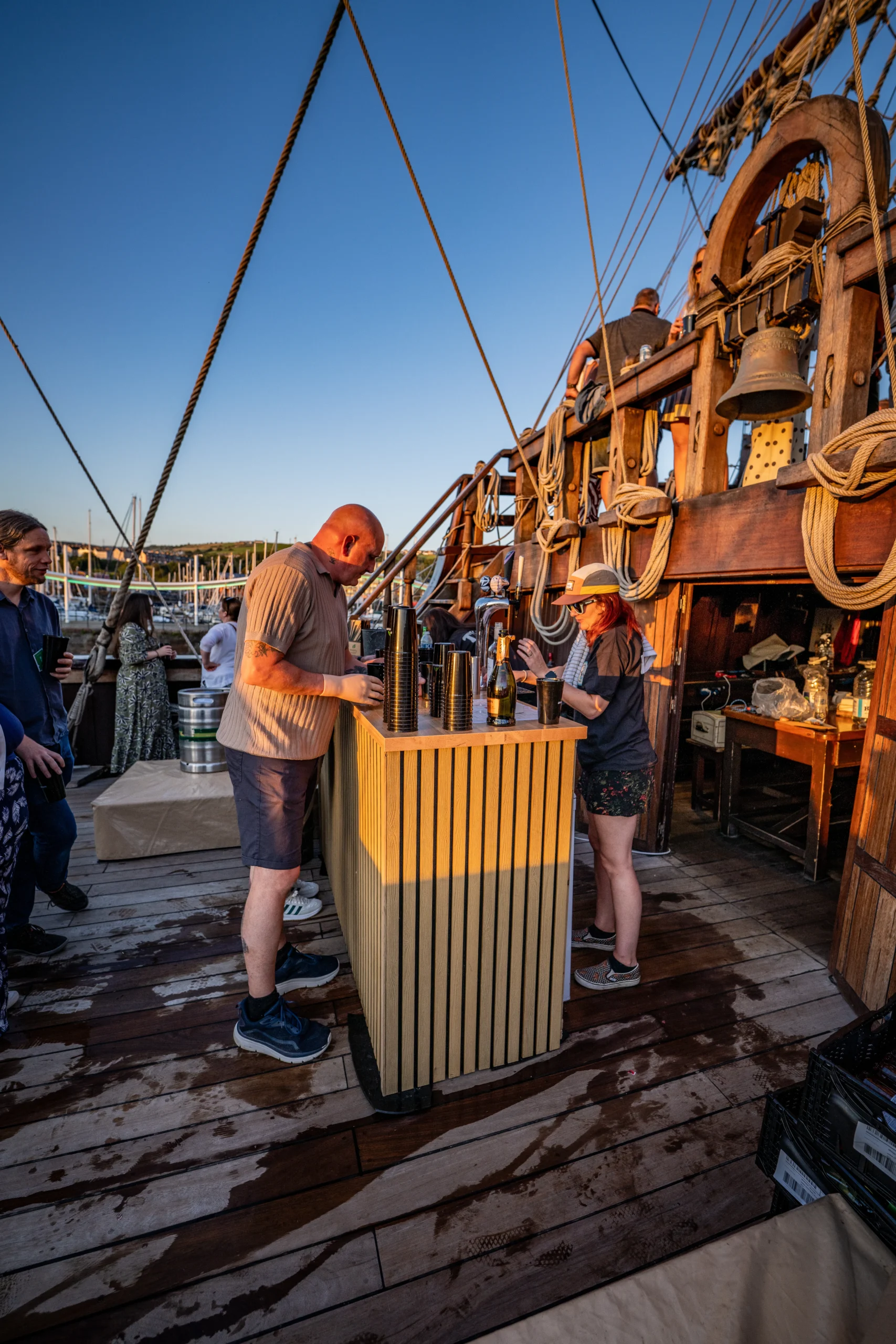 Two people serving drinks at an outdoor bar setup on a wooden ship deck