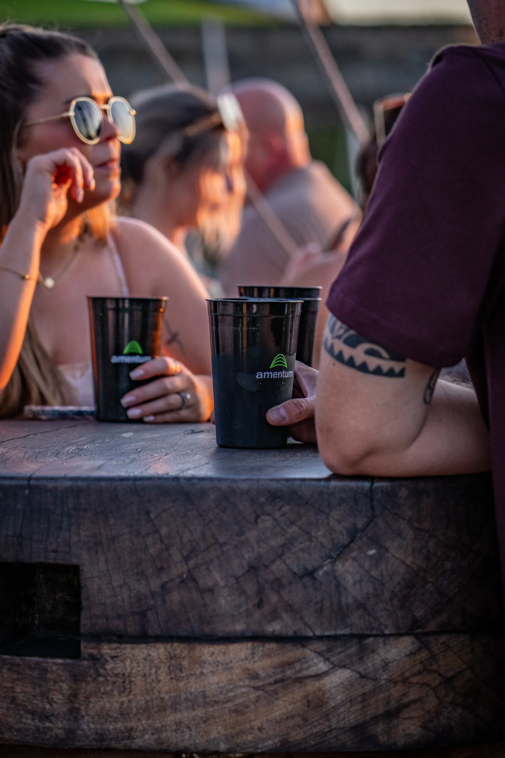 People socialising outdoors at a wooden table, holding branded reusable cups at sunset,