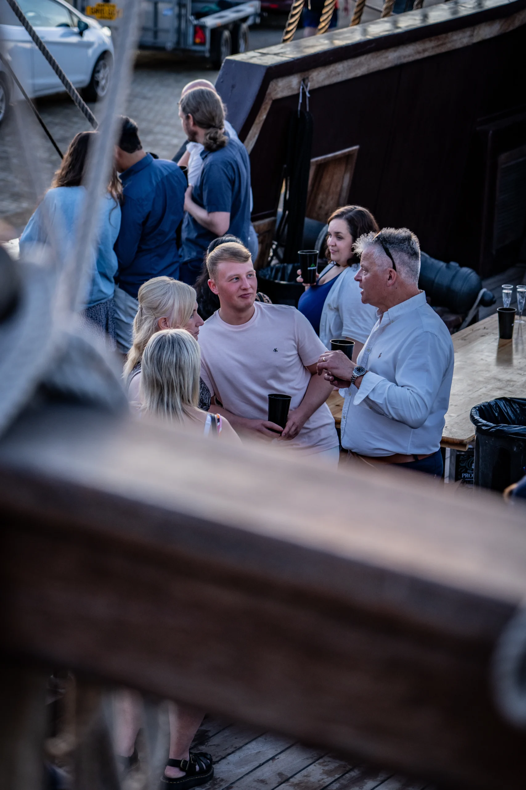 Group of people socialising on the deck of a wooden ship during an event, holding drinks and talking casually.