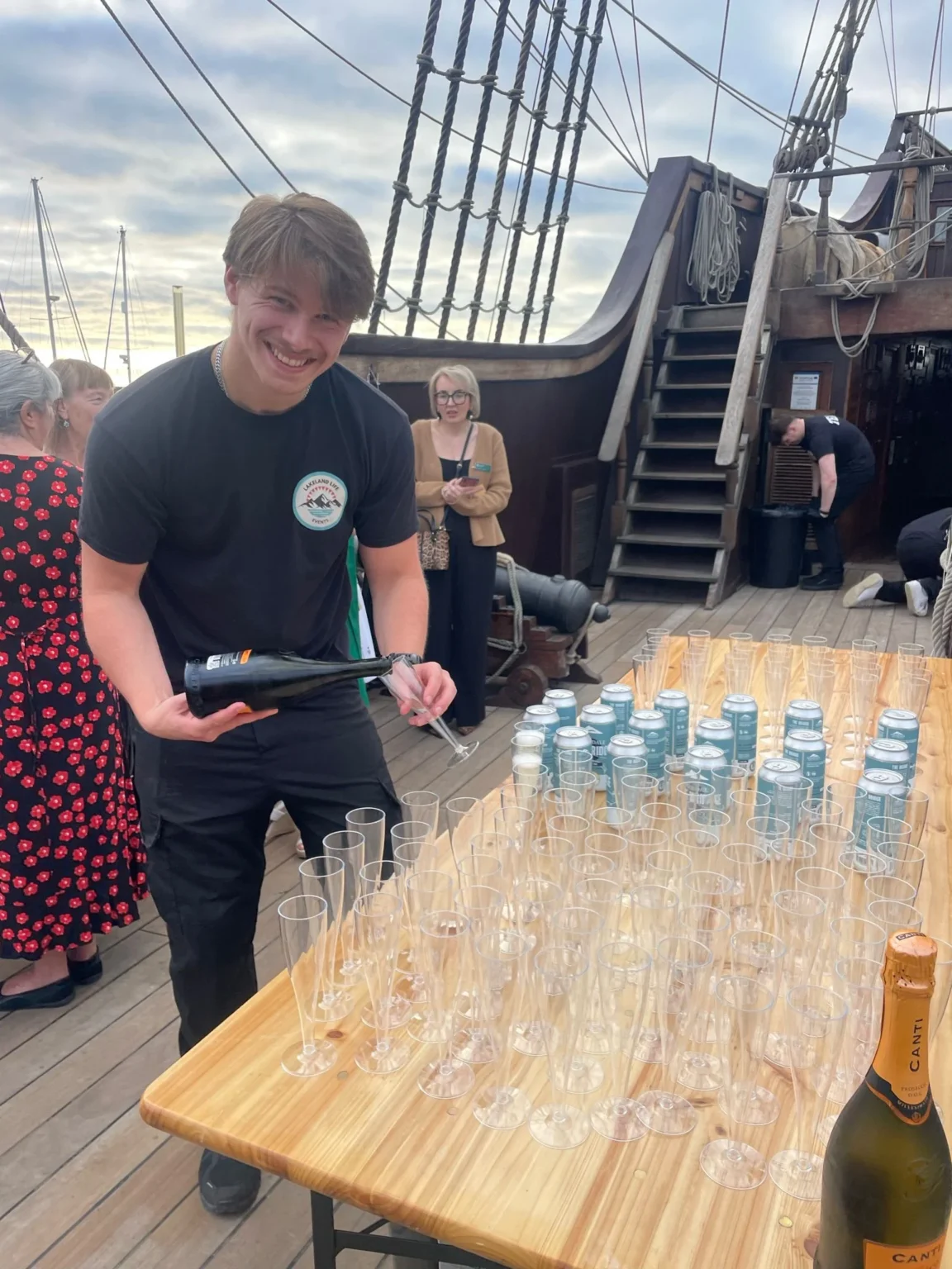 Smiling event staff member pouring drinks at a table of empty glasses on a historic wooden ship.
