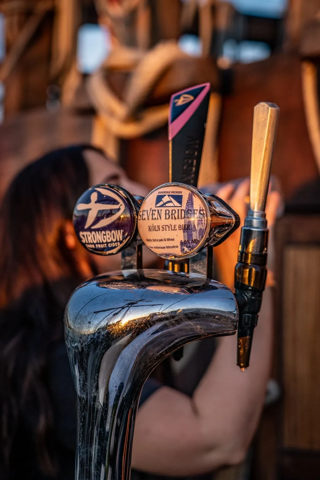 Close-up of beer taps labeled Strongbow and Seven Bridges with a person pouring a drink in the background.