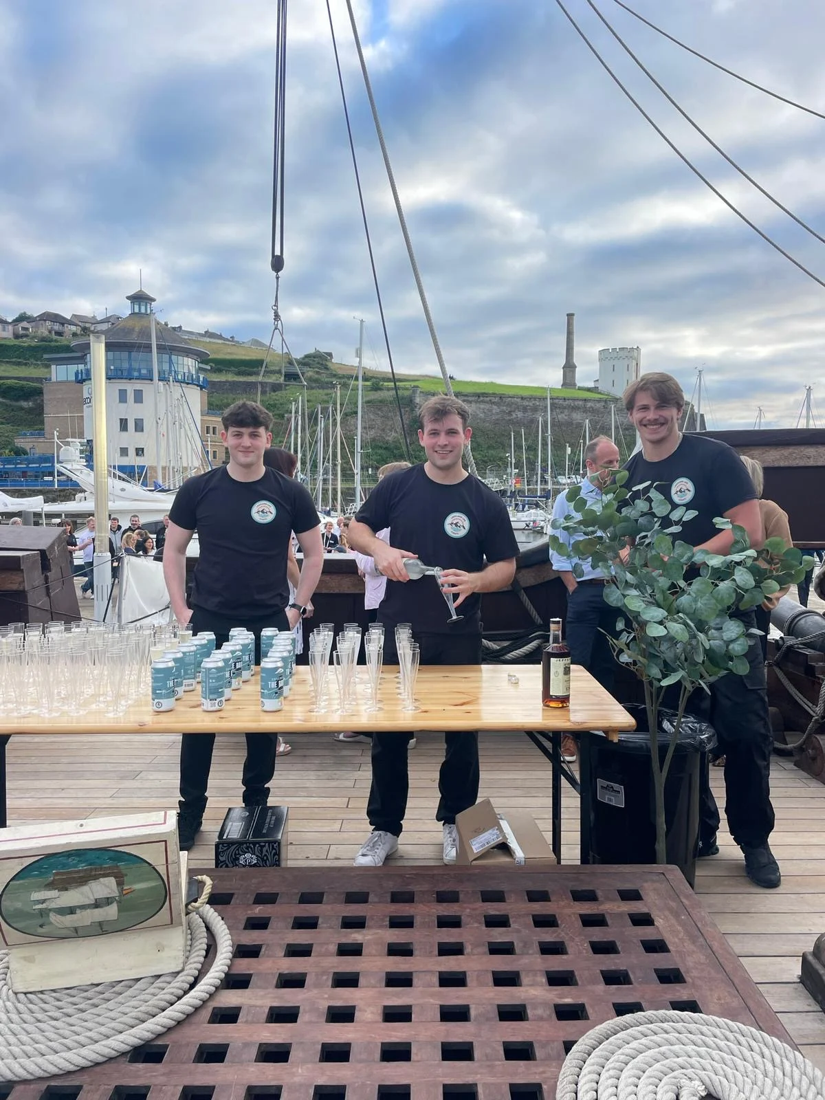 Table on a wooden ship deck set with rows of empty plastic glasses and canned drinks, with staff working the bar.