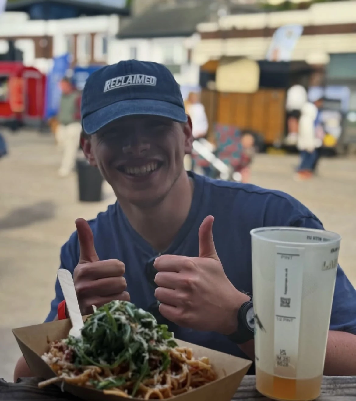 A smiling young man wearing a blue “Reclaimed” cap gives two thumbs up while sitting at an outdoor food event, with a bowl of pasta and a large drink cup in front of him.