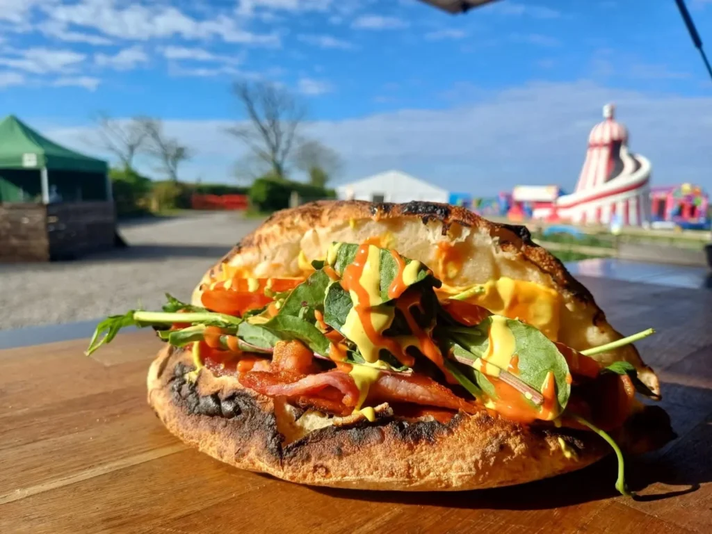 A toasted sandwich filled with bacon, fresh spinach, and sauces, resting on a wooden counter outdoors with a sunny festival scene and funfair rides blurred in the background.