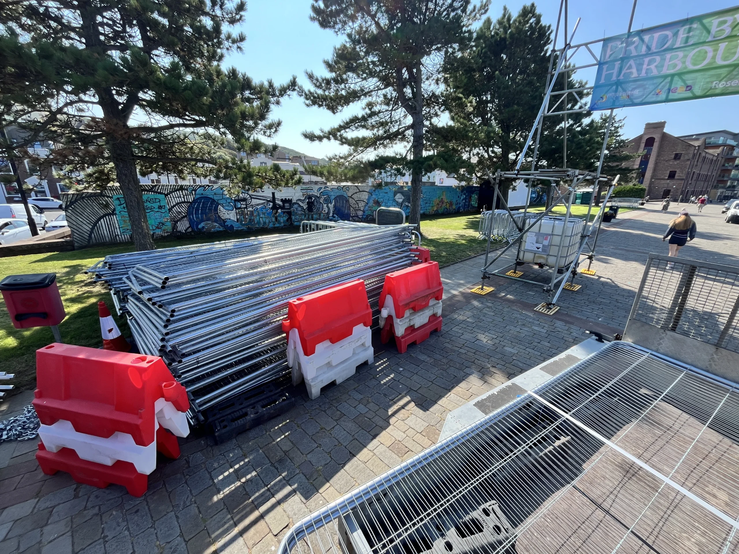Stacks of metal crowd-control barriers and red-and-white plastic barricades arranged on a paved area near trees, with event equipment and scaffolding visible in the background.
