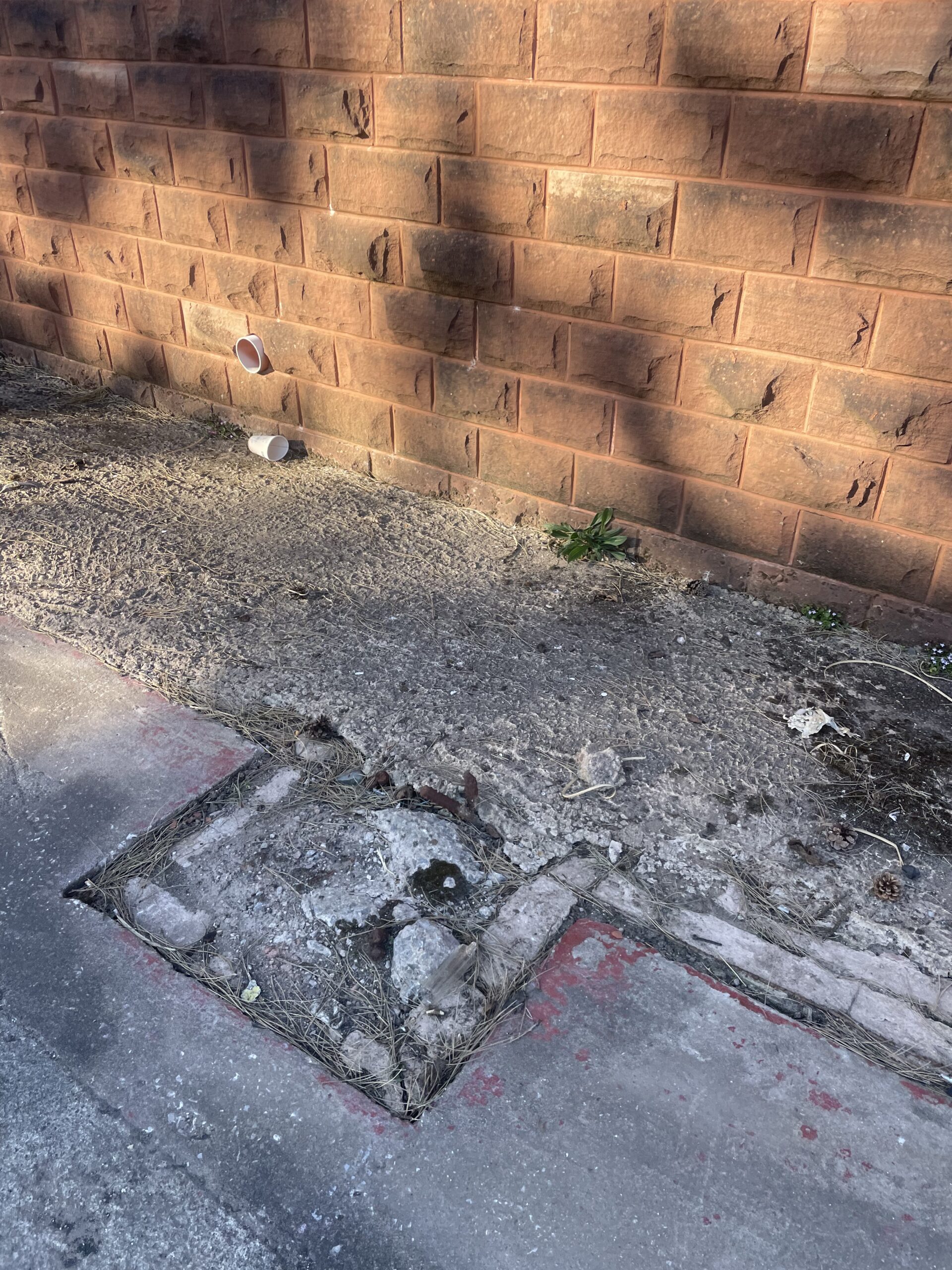 An area of concrete that has been cut out beside a brick wall, showing a square section of removed pavement where work has been done for event setup.