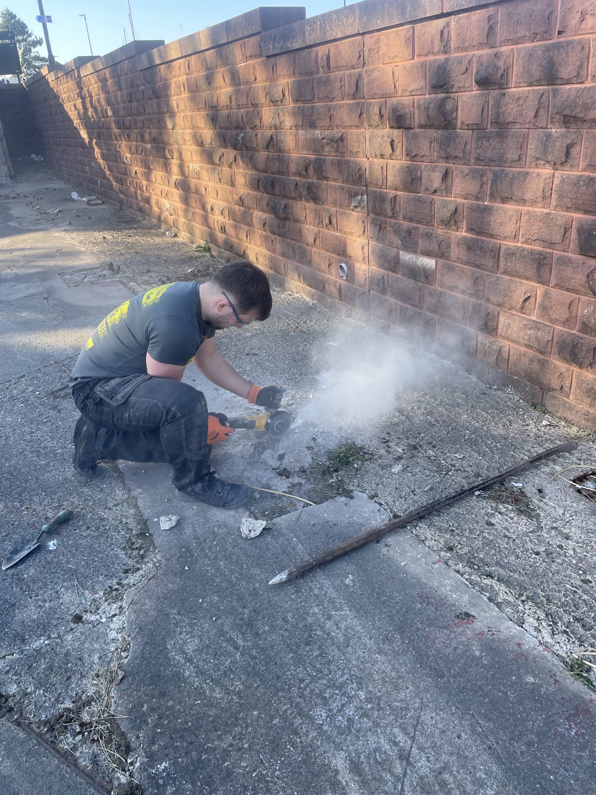 Person kneeling on the ground using an angle grinder to cut or break up old concrete next to a brick wall, with dust rising from the work