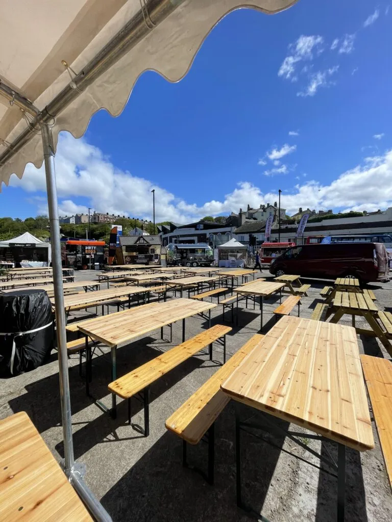 Outdoor event setup with rows of wooden tables and benches under a canopy on a sunny day.